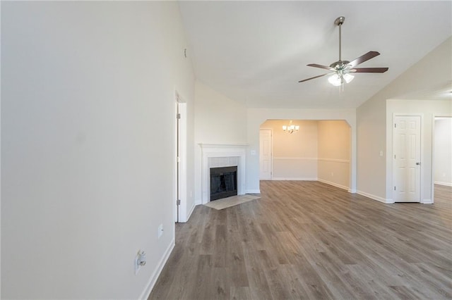 unfurnished living room with ceiling fan with notable chandelier, vaulted ceiling, a tiled fireplace, and wood finished floors