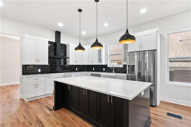 kitchen with wall chimney range hood, a kitchen island, stainless steel refrigerator with ice dispenser, white cabinets, and light wood-type flooring