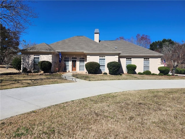 ranch-style home with stucco siding, roof with shingles, a chimney, and a front lawn