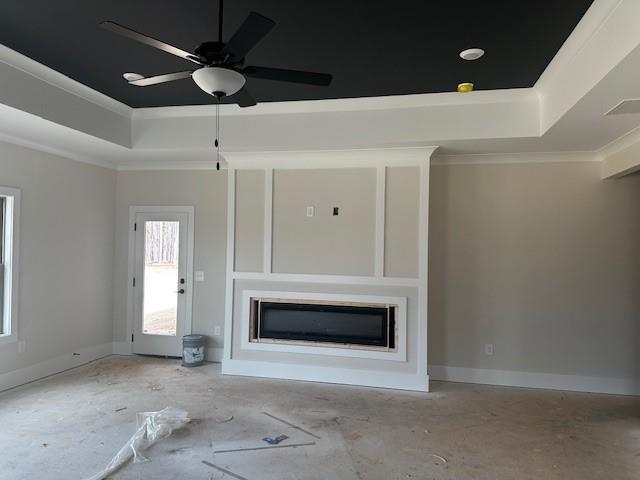 unfurnished living room featuring baseboards, a ceiling fan, a glass covered fireplace, ornamental molding, and a tray ceiling