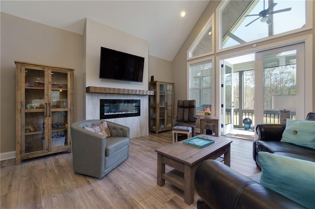 living area featuring baseboards, wood finished floors, a glass covered fireplace, high vaulted ceiling, and a ceiling fan