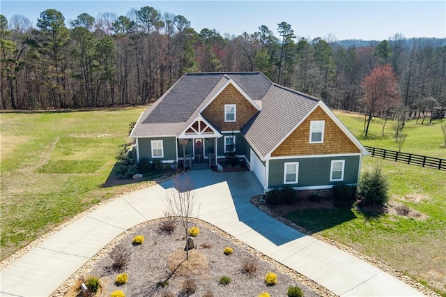 craftsman-style house featuring a shingled roof, a forest view, fence, a front yard, and driveway