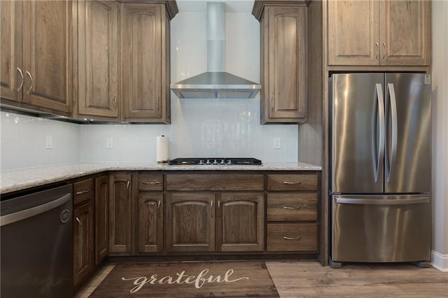 kitchen with light stone countertops, wood finished floors, decorative backsplash, appliances with stainless steel finishes, and wall chimney exhaust hood