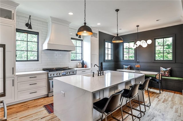 kitchen featuring a kitchen island with sink, white cabinets, custom range hood, light wood finished floors, and stainless steel range