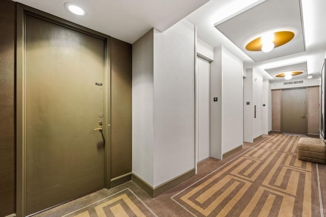 hallway with elevator, light colored carpet, visible vents, and baseboards