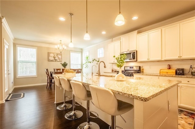 kitchen featuring appliances with stainless steel finishes, a kitchen island with sink, hanging light fixtures, white cabinetry, and ornamental molding