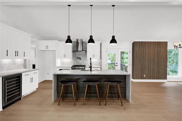 kitchen with white cabinets, an island with sink, wall chimney range hood, and beverage cooler