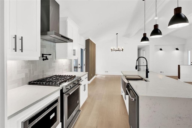 kitchen featuring wall chimney range hood, appliances with stainless steel finishes, white cabinetry, hanging light fixtures, and an island with sink