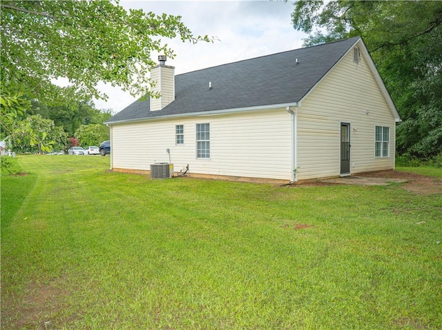 back of house featuring a shingled roof, a yard, a chimney, and central AC unit