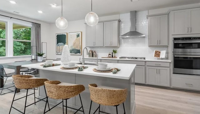 kitchen with appliances with stainless steel finishes, wall chimney exhaust hood, sink, gray cabinets, and an island with sink