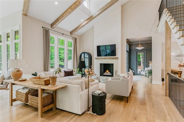 living room with beam ceiling, a lit fireplace, high vaulted ceiling, stairway, and light wood-style flooring