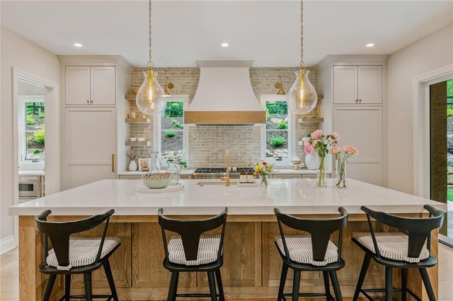 kitchen featuring a healthy amount of sunlight, backsplash, a large island with sink, light countertops, and custom range hood