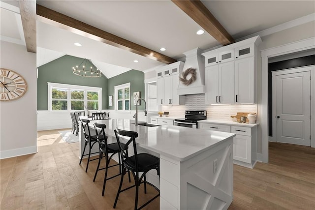 kitchen with light wood finished floors, custom exhaust hood, a sink, a kitchen island with sink, and stainless steel electric range oven