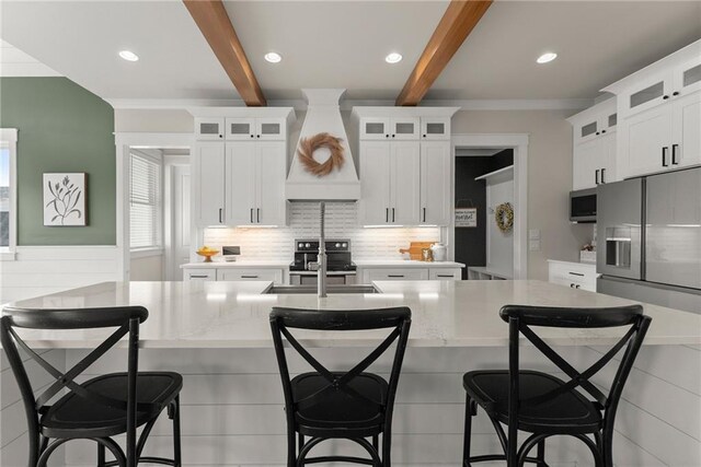 kitchen featuring a sink, decorative backsplash, a kitchen island with sink, premium range hood, and beam ceiling