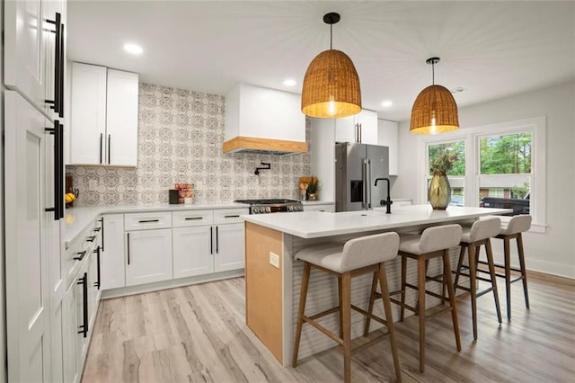 kitchen with an island with sink, custom exhaust hood, stainless steel fridge with ice dispenser, light countertops, and light wood finished floors