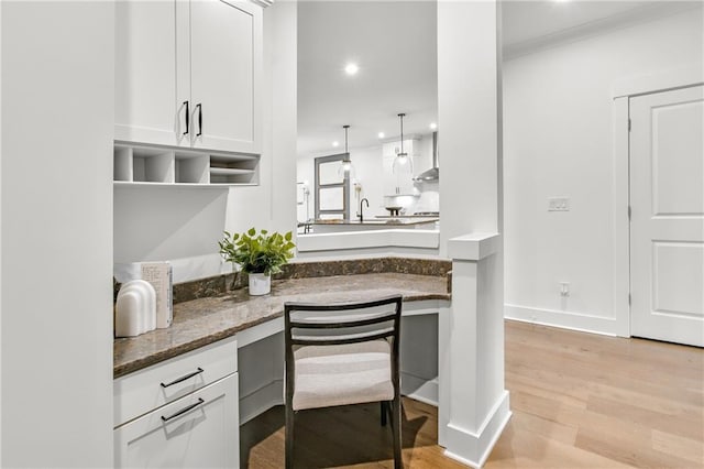 kitchen featuring decorative light fixtures, wall chimney range hood, light wood-type flooring, white cabinetry, and dark stone counters