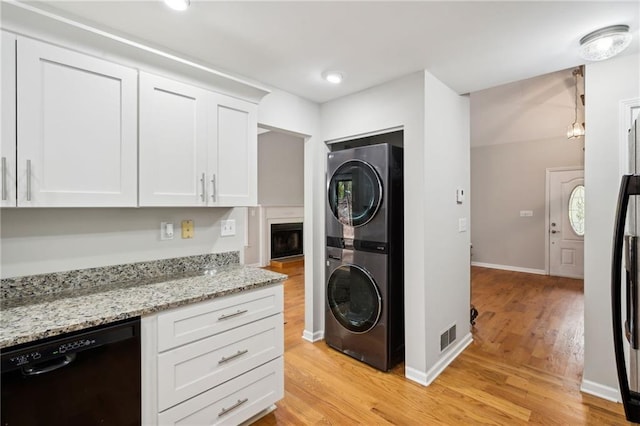 kitchen featuring light stone counters, light wood-style flooring, stacked washer / dryer, white cabinetry, and black dishwasher