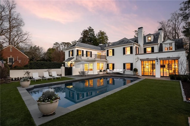 back house at dusk with a fenced in pool, an outdoor hangout area, a balcony, and a lawn