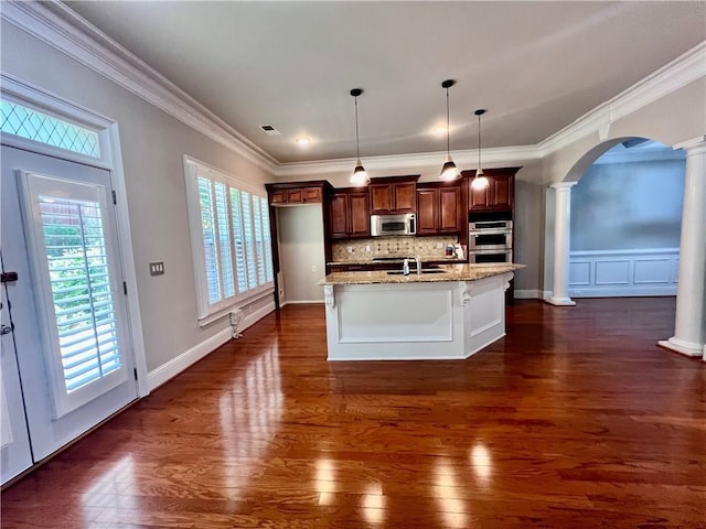 kitchen featuring appliances with stainless steel finishes, pendant lighting, decorative columns, light stone countertops, and a center island with sink