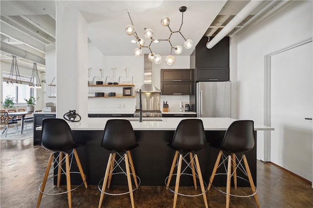 kitchen featuring an inviting chandelier, a kitchen breakfast bar, wall chimney exhaust hood, stainless steel fridge, and an island with sink