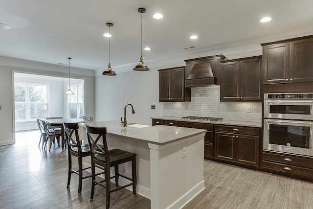 kitchen featuring premium range hood, a sink, backsplash, light wood-style floors, and appliances with stainless steel finishes