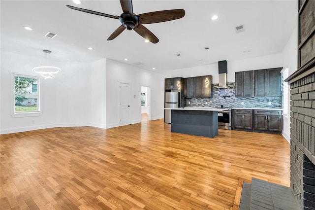 kitchen featuring decorative light fixtures, wall chimney range hood, appliances with stainless steel finishes, a center island, and ceiling fan with notable chandelier