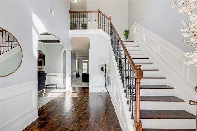 stairway with a towering ceiling, a decorative wall, hardwood / wood-style floors, arched walkways, and a wainscoted wall