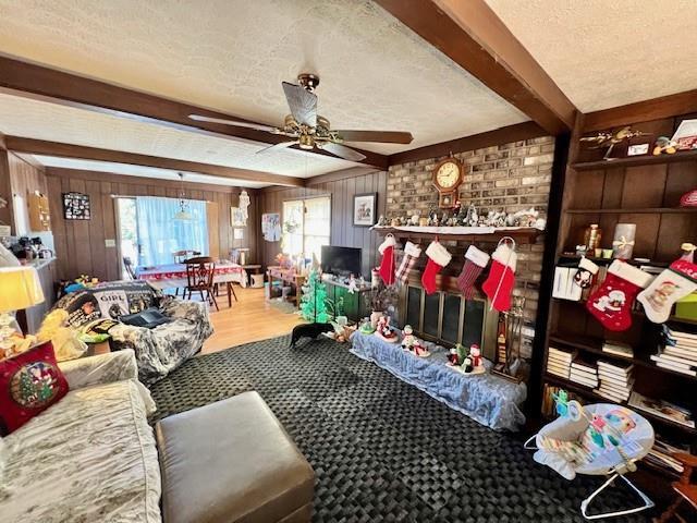 living room featuring wooden walls, a ceiling fan, beamed ceiling, a textured ceiling, and a brick fireplace