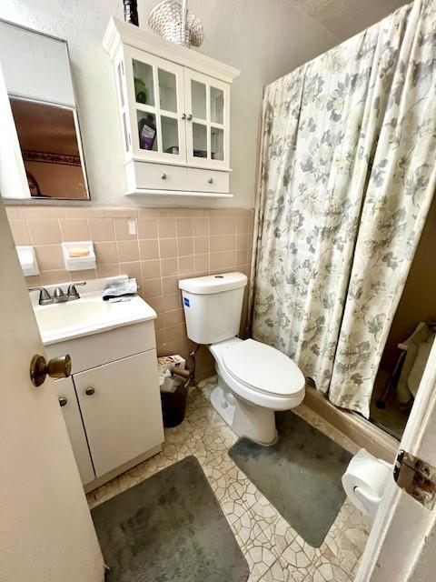 full bath featuring toilet, vanity, wainscoting, a stall shower, and tile walls