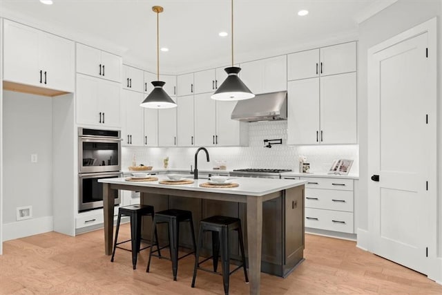 kitchen with white cabinetry, appliances with stainless steel finishes, light wood-type flooring, an island with sink, and wall chimney exhaust hood