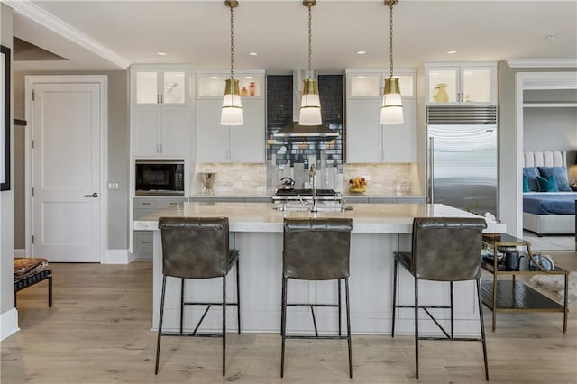 kitchen with wall chimney range hood, built in appliances, an island with sink, white cabinetry, and backsplash