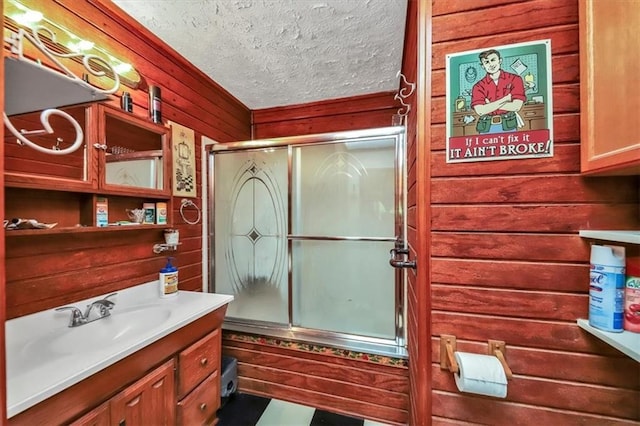 bathroom with a textured ceiling, combined bath / shower with glass door, wood walls, and vanity