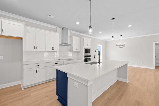 kitchen featuring wall chimney exhaust hood, decorative light fixtures, white cabinetry, sink, and a center island with sink