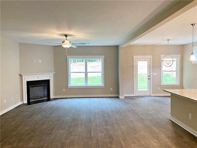 unfurnished living room with baseboards, a fireplace with flush hearth, dark wood-style floors, and ceiling fan with notable chandelier