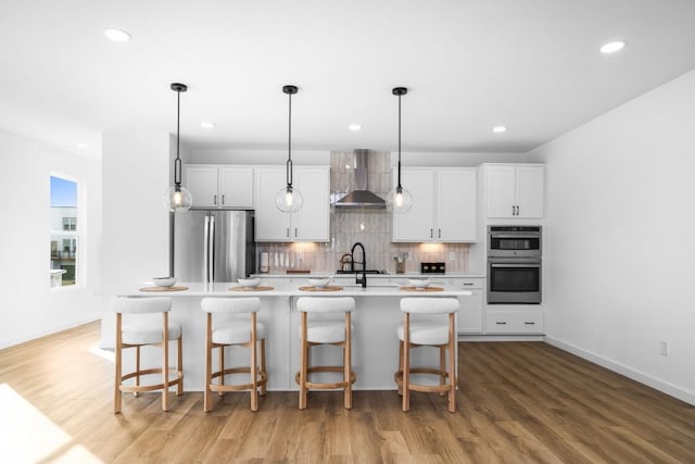 kitchen featuring wall chimney exhaust hood, a breakfast bar, a center island with sink, stainless steel appliances, and white cabinets