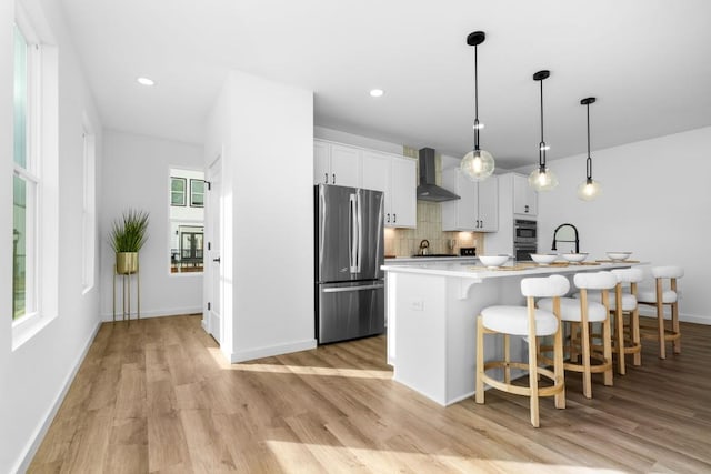 kitchen featuring pendant lighting, wall chimney range hood, appliances with stainless steel finishes, white cabinets, and a center island with sink