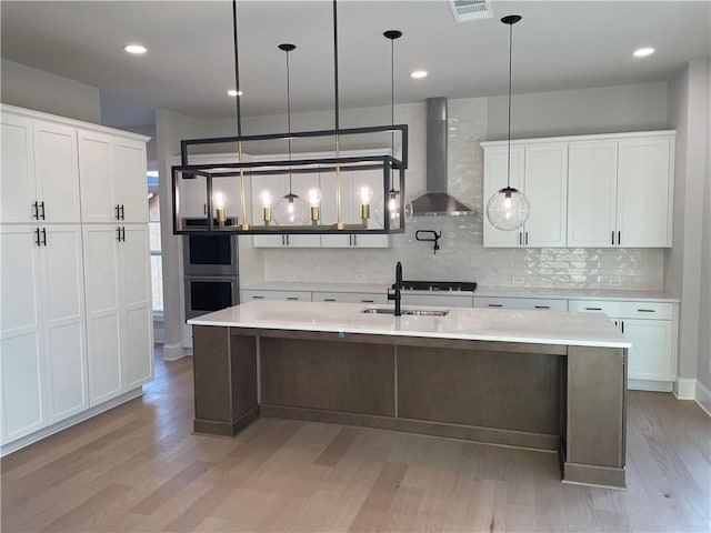 kitchen with wall chimney range hood, sink, white cabinetry, a center island with sink, and decorative light fixtures