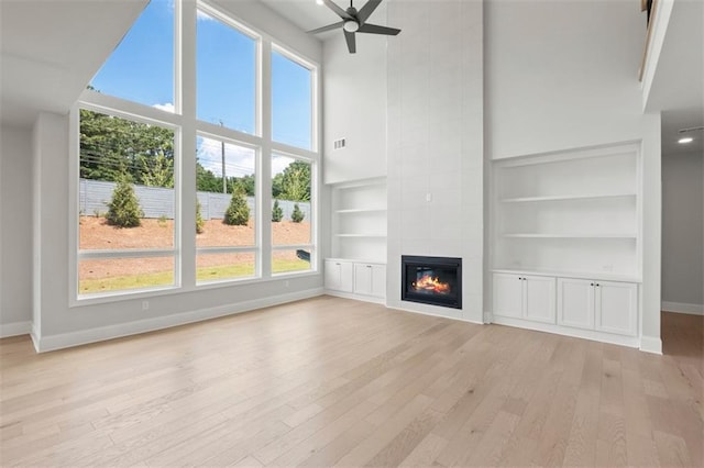 unfurnished living room featuring a towering ceiling, built in features, a fireplace, ceiling fan, and light wood-type flooring