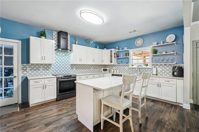 kitchen with a breakfast bar, stainless steel electric range oven, white cabinetry, wall chimney exhaust hood, and open shelves