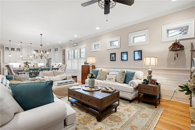 living room featuring light wood finished floors, crown molding, a wainscoted wall, ceiling fan with notable chandelier, and recessed lighting