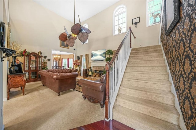 staircase featuring ceiling fan with notable chandelier, wood finished floors, carpet, a brick fireplace, and a towering ceiling