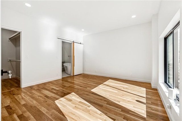 unfurnished bedroom featuring wood finished floors, a barn door, baseboards, recessed lighting, and a walk in closet
