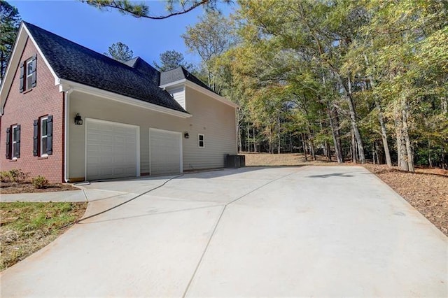 view of side of property featuring a shingled roof, brick siding, a garage, cooling unit, and concrete driveway