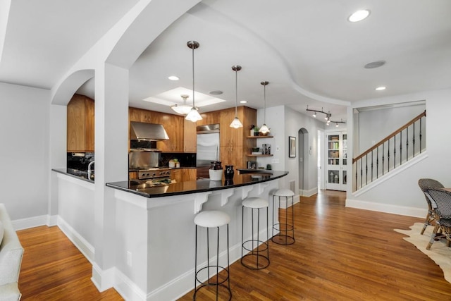 kitchen featuring wood finished floors, arched walkways, built in refrigerator, wall chimney exhaust hood, and range