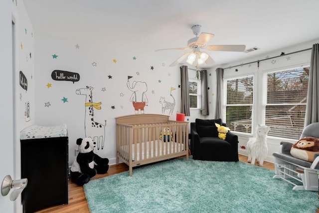 bedroom featuring visible vents, a nursery area, a ceiling fan, and light wood finished floors