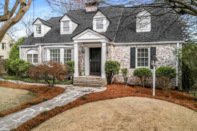 cape cod home featuring brick siding, a chimney, and a shingled roof