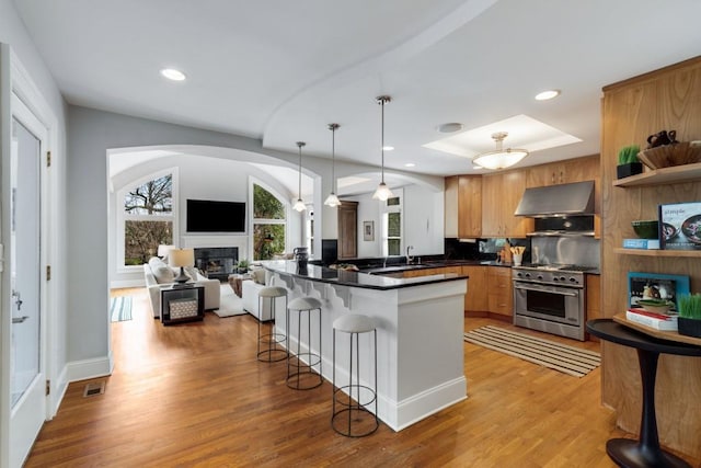 kitchen with open shelves, under cabinet range hood, high end stainless steel range, a glass covered fireplace, and a kitchen breakfast bar