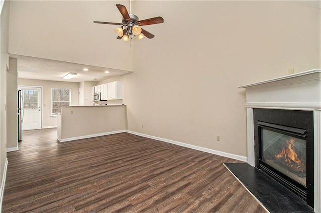 unfurnished living room with dark wood finished floors, a towering ceiling, a glass covered fireplace, ceiling fan, and baseboards