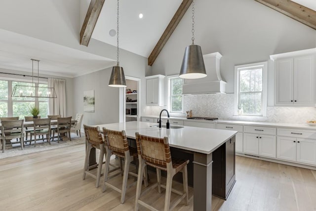 4781 Falling Brook Dr, Flowery Branch, GA 30542 - kitchen featuring wall chimney range hood, light wood-style flooring, an island with sink, beamed ceiling, and tasteful backsplash