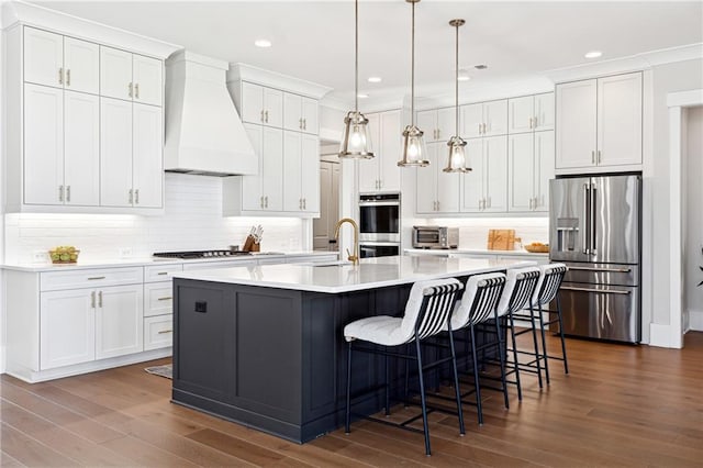 kitchen with custom exhaust hood, appliances with stainless steel finishes, white cabinetry, hanging light fixtures, and a center island with sink